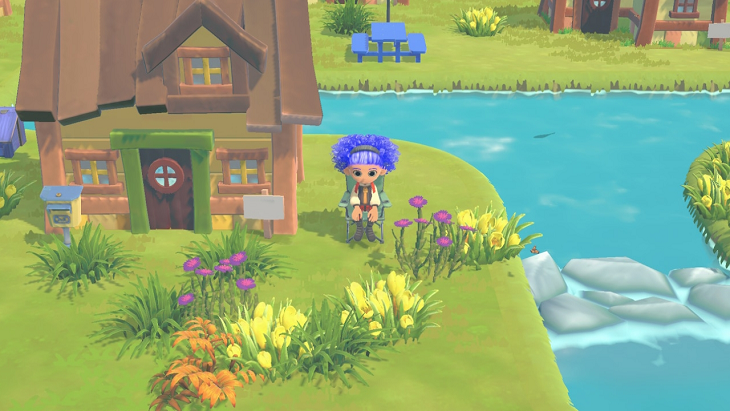 Game's avatar sits in an outdoor camping chair next to a small yellow cottage and a rocky bridge that crosses over a blue river. There are yellow and brown plants in the yard.